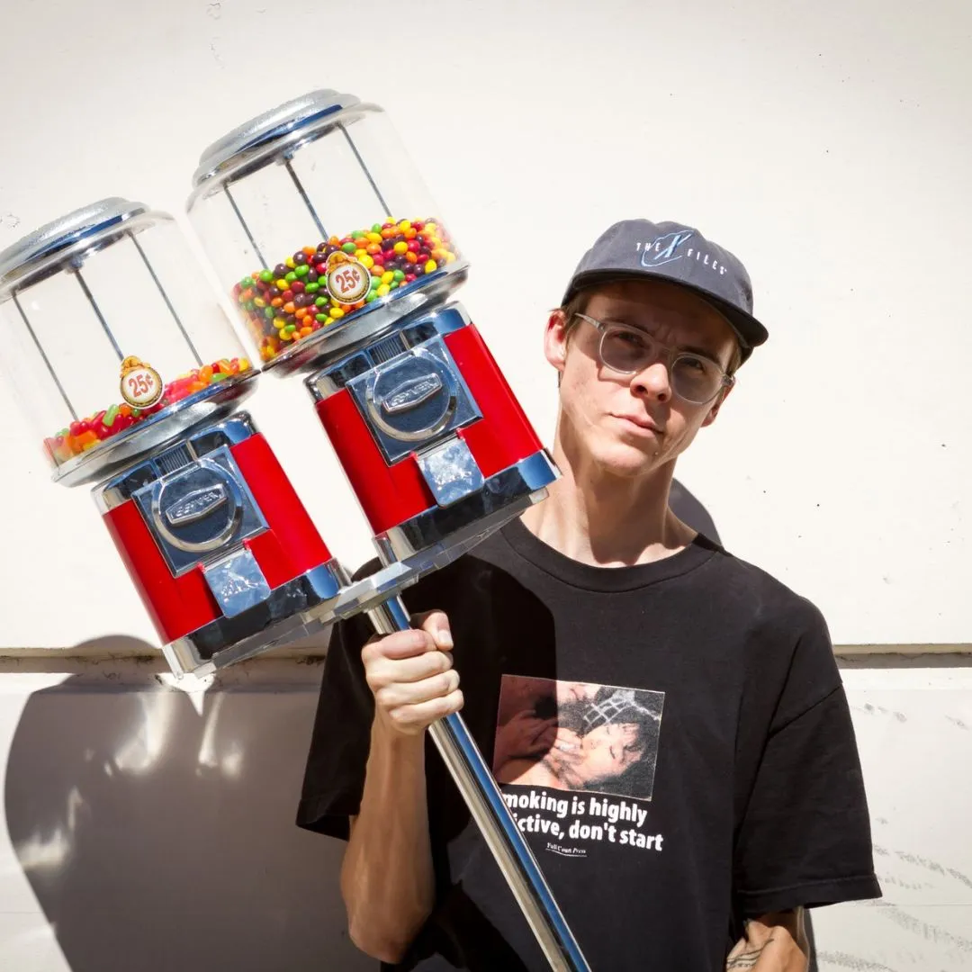 a headshot of Side Hustle Art Show artist Garrett Moore wearing a black shirt, grey hat, and holding a red candy machine