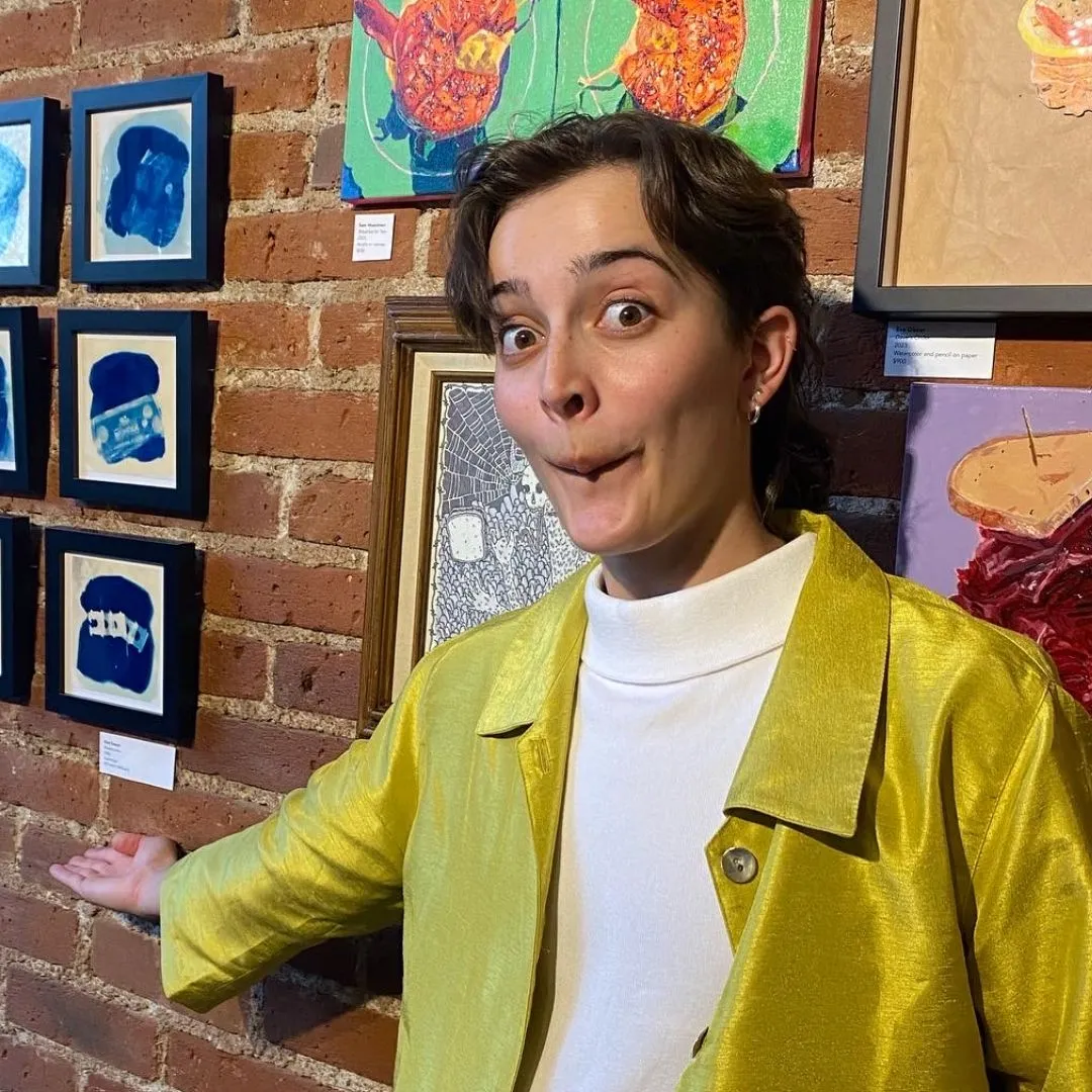 a headshot of Side Hustle Art Show artist Kate Sawyer wearing a white shirt and a yellow coat in front of a brick wall with art hanging on it