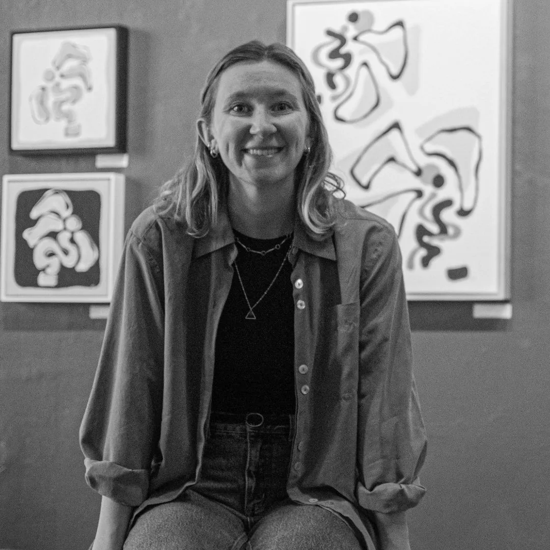 Headshot of Side Hustle Art Show artist, Rah Bottini sitting in front of some of her paintings handing on a wall